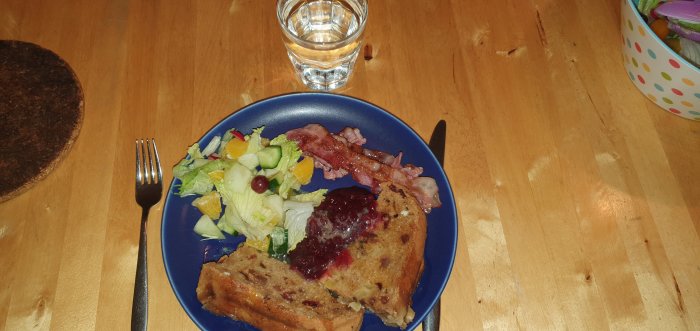 Poffert med lingon och smält smör, stekt bacon och sallad på blå tallrik med vattenglas.