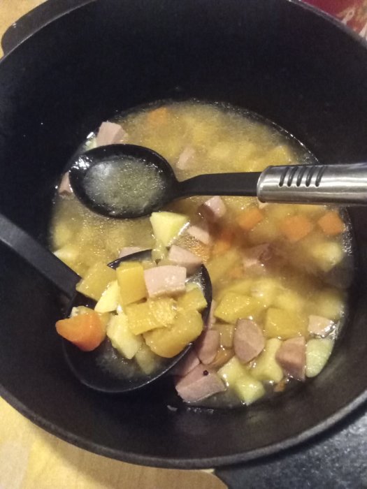 En gryta med korv och tärnade rotfrukter, lagad i en svart gjutjärnsgryta med slev.