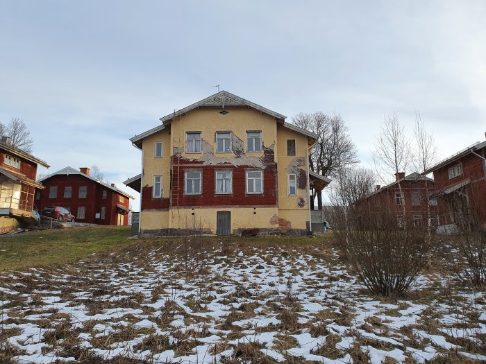Gammalt gulaktigt hus med röda detaljer från 1896 på en vintergräsmatta med snöfläckar.