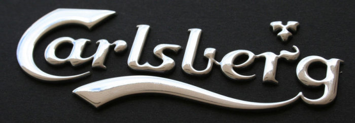 Silverfärgad Carlsberg-logotyp i metall mot svart bakgrund.