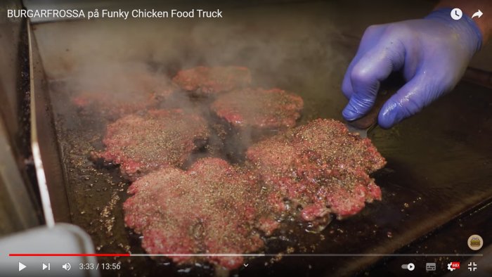 Hamburgare med riklig peppar på stekbord, rök och en hand i blå handske vid en kant.