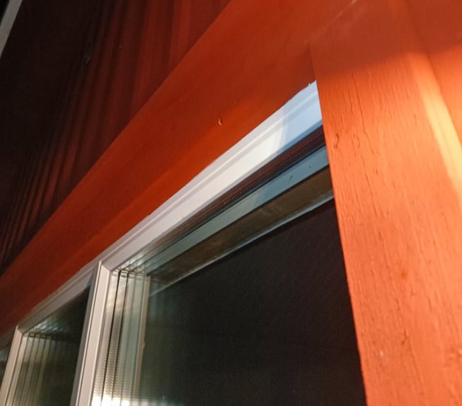 Närbeläget fönster med ventilspringa synlig under takutsprånget i skyddat läge, omgiven av röda foderelement.