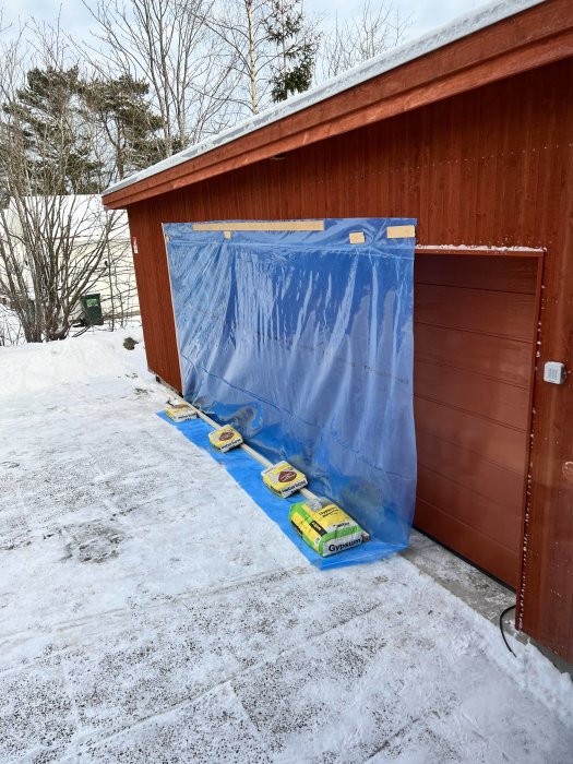 Plastskydd för byggprojekt täcker en del av en röd byggnad och mark, säckar med byggmaterial väger ner skyddet.