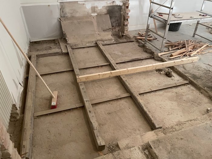 Upprivet gammalt plankgolv och exponerat betonggolv under renovering med synliga reglar och nivåskillnader.