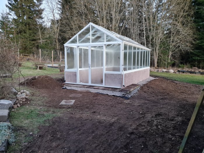 Nytt växthus med klar glasbeklädnad och rosa fundament omgivet av nyanlagd jord och trädgårdsarbete.