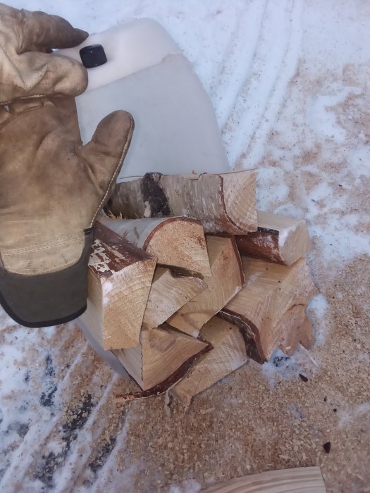 Handskeklädd hand som visar klyvna björkvedsklumpar och sågspån på snöig mark.