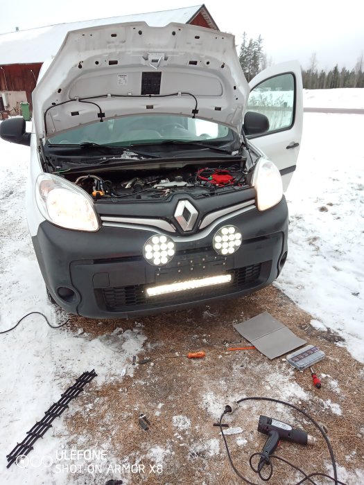 Bil med öppen huva där en extraljusramp med blixtljus monterats, verktyg syns på marken.