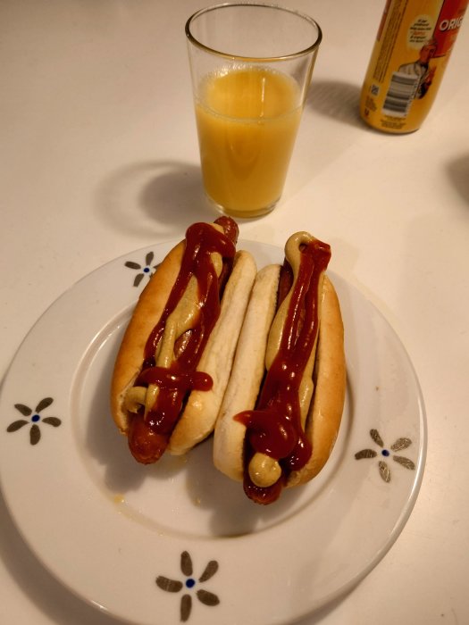 Två korvar med bröd och ketchup på tallrik bredvid ett glas apelsinjuice och en juicekartong.