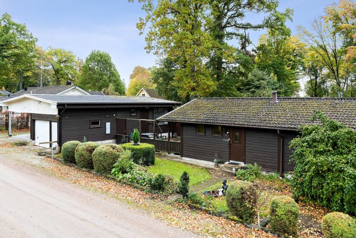 Bild på ett enfamiljshus med brun panel, tegeltak och garage, omgivet av en trädgård och lövfällande träd.