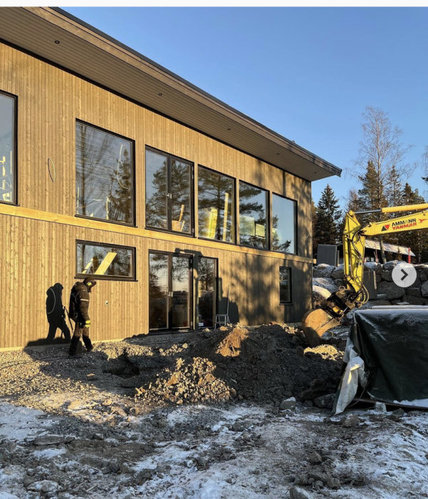 Modern suterränghus under konstruktion med grävmaskin och röran av byggmaterial framför.