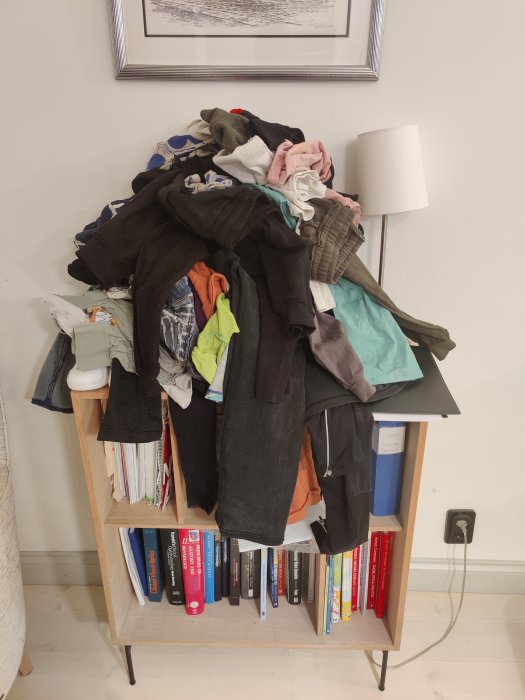 Bokhylla med böcker täckt av kläder och en tvättkorg ovanpå i ett hem.