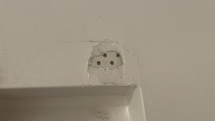 Skador på vit vägg där målarfärg lossnat och avslöjar grått material och fyra små hål.