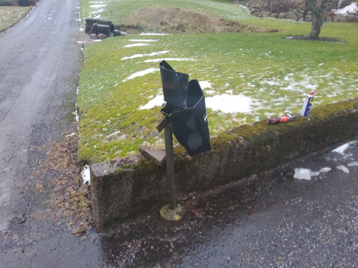 Skadad svart brevlåda som hänger på sned på en stolpe, vid en mur med snöfläckar på marken.