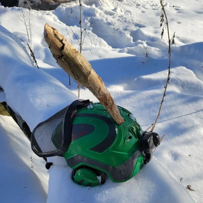 Säkerhetshjälm med visir i snön med en stor trädstam som fallit ovanpå.