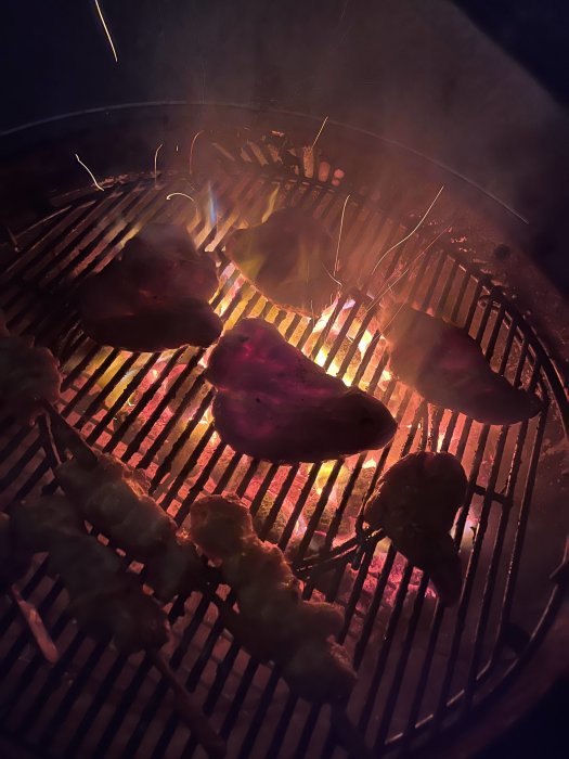 Kvällsbelysning över en grill med biffar och spett som grillas över glödande kol.