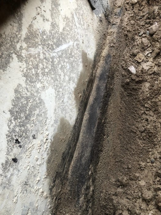 Grund av ett hus med blottlagda betongväggar och en 8 cm tjock betongklack synlig i en grävd dräneringsränna.
