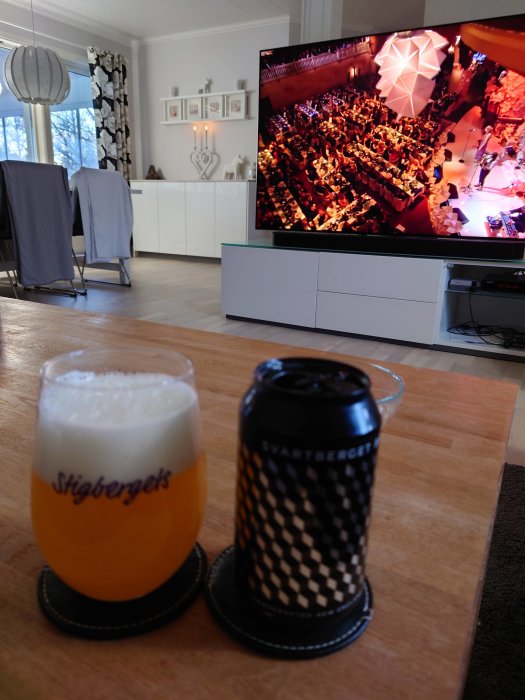 Ölglas med logotyp och en ölburk på ett bord framför en TV som visar en konsert.