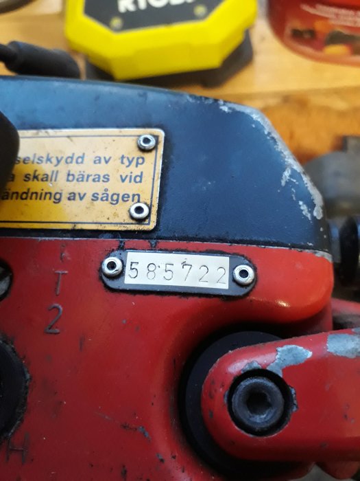 Del av röd maskin med tydlig serienummerplåt som visar numret 585722 och text på gul etikett om öronskydd.