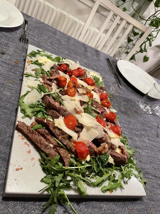 En serverad biff tagliata med rucola, parmesan och rostade tomater på ett vitt serveringsfat.