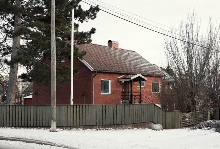 Rött tegelhus från 40-talet med svart tegeltak och grönt staket, omgivet av träd och snö.