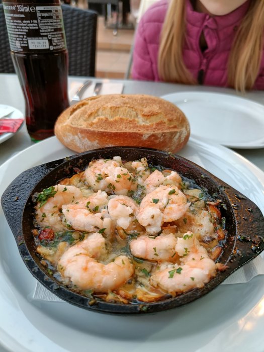 Gambas al ajillo i gjutjärnsstekpanna med tillbehör av bröd och läsk på restaurangbord.
