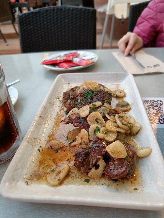 Grillat kött toppat med vitlöksklyftor och örter på ett vitt serveringsfat, med en dryck och en person i bakgrunden.