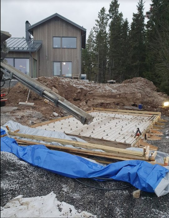 Nyligen gjuten platta för utekök med träramar och armeringsjärn, omgivet av en grushög och ett hus i bakgrunden.