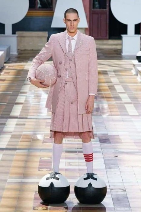Manlig modell går på catwalk klädd i rosa randig kostym med kjol, håller i matchande hatt, står på svartvita bollar.