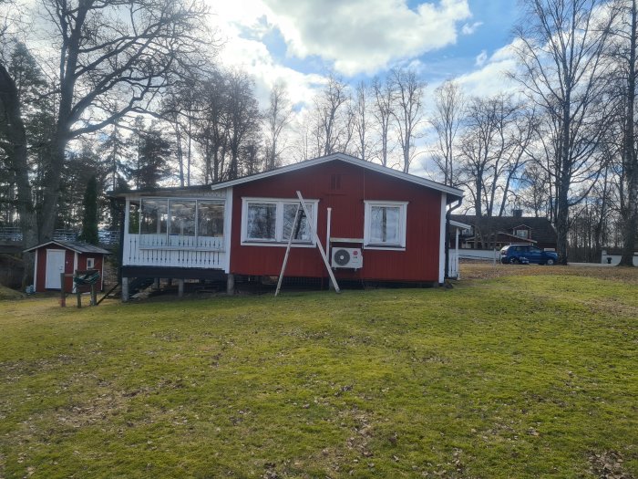 Röd stuga med vit knut och veranda, belägen på en gräsmatta med träd i bakgrunden.