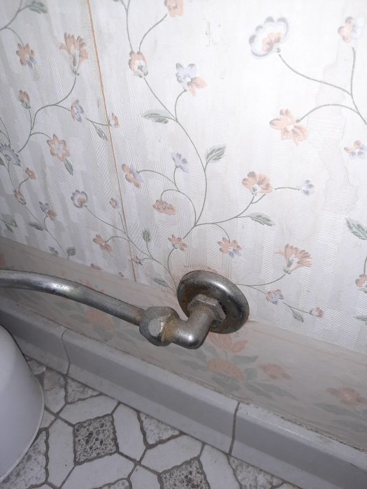 Gammalt rostigt vattenrör med metallkoppling vid blommigt tapetserad vägg och kaklat golv.