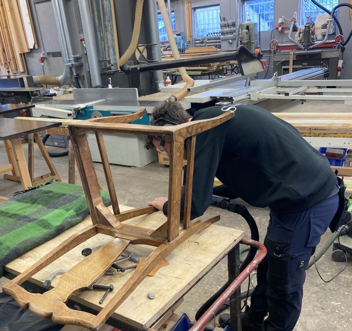 Person som reparerar en stol i ett snickeri med verktyg och träbearbetningsmaskiner i bakgrunden.