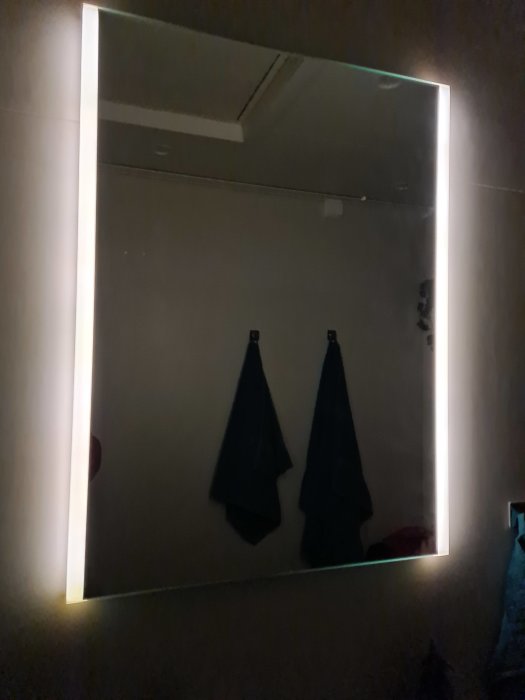 Badrumsspegel med tända sidolampor som ger svagt ljus, handdukar hänger under spegeln.