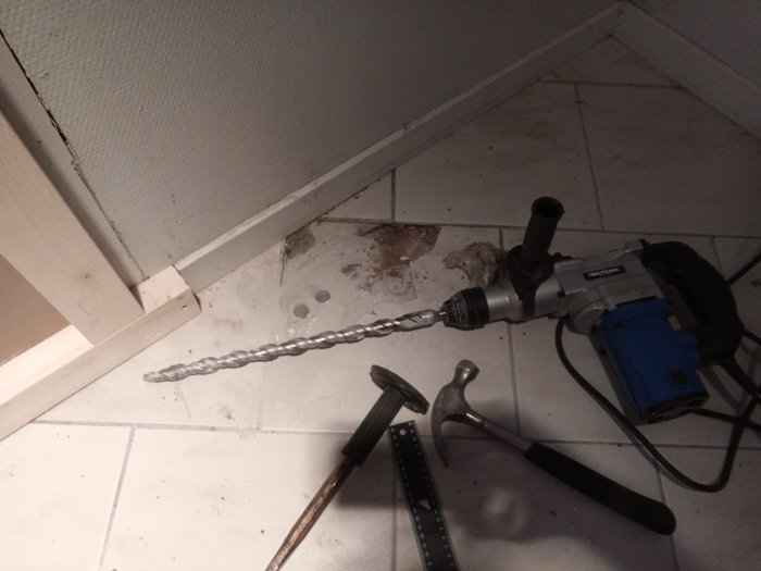 Borrmaskin och borr på ett stenbelagt golv med borrade hål i en betongvägg, tillsammans med hammare och måttband.