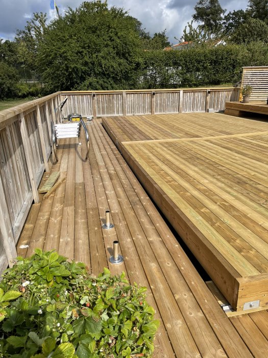 Trädäck i trädgård med räcke, inbyggd trappa och plantering, verktyg på bänk under solig himmel.