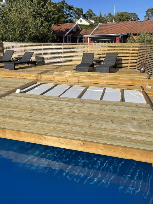 Trädäck med solstolar och integrerad pool framför röda hus och gröna träd i dagsljus.