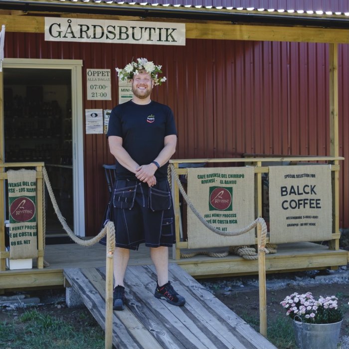 Leende man i svart t-shirt och svart kjol står framför en gårdsbutik med kaffesäckar.