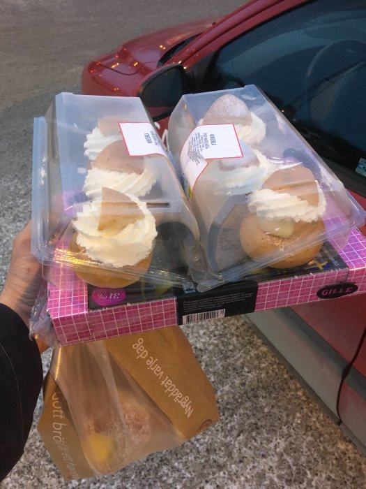 Hand håller förpackningar med semlor vid en parkerad bil i skymningen.