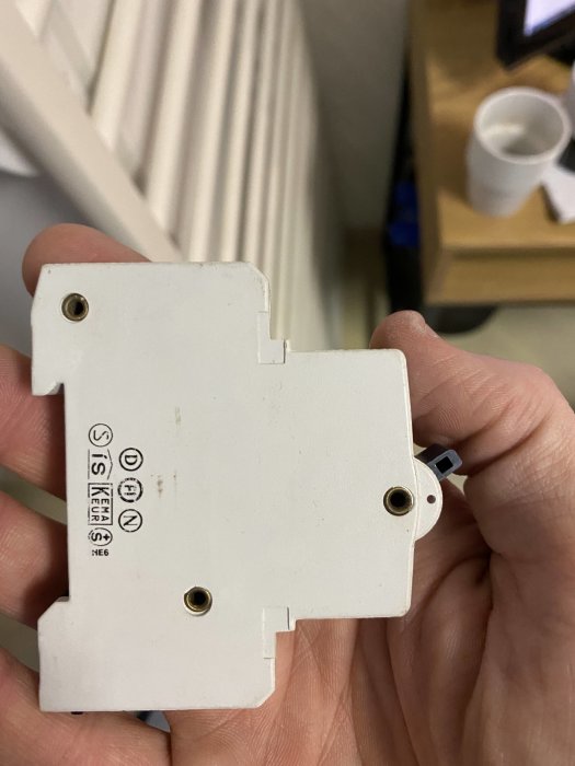 Hand håller vit elektrisk installationskomponent med märkningar och skruvhål.