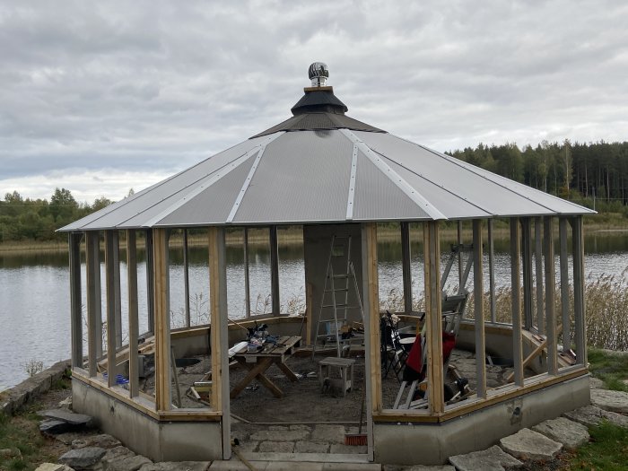 Okonstruerad träpaviljong med plåttak vid en sjö under uppbyggnad, byggmaterial och verktyg syns.