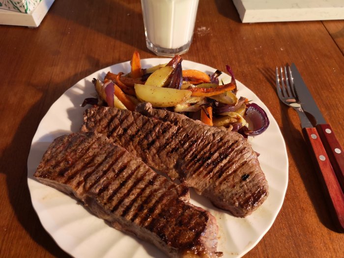Grillade biffar med rostade grönsaker på tallrik bredvid ett glas mjölk.