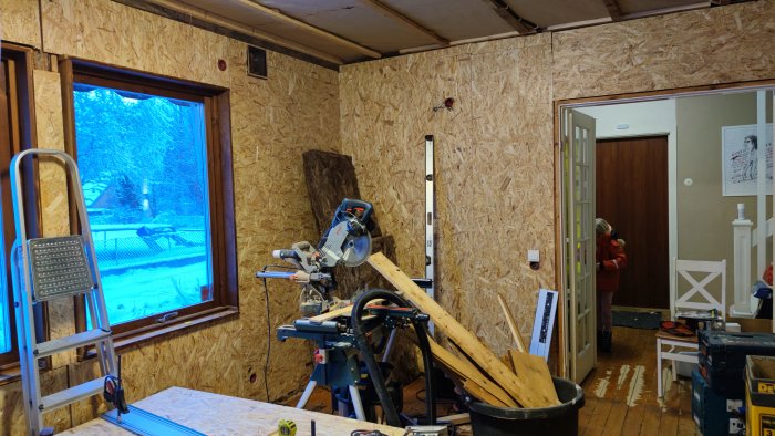 Renoveringsarbete med OSB-skivor på väggar, arbetsverktyg och snötäckt landskap synligt genom fönstret.