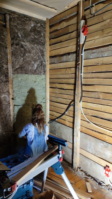 Person som arbetar på en vägg under renovering med synlig isolering och eldragning, framför en arbetsbänk med verktyg.
