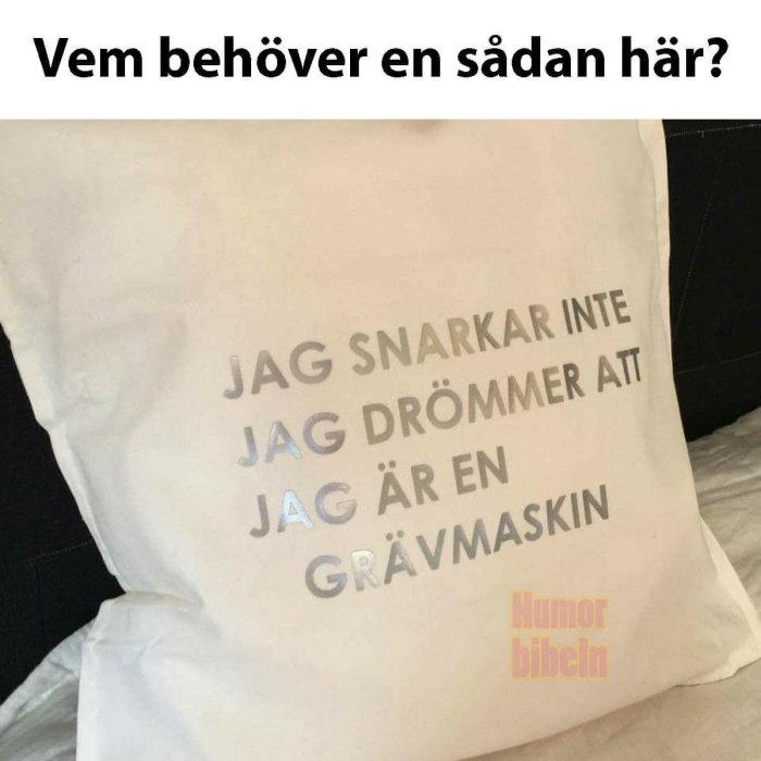 Vit kudde med text "Jag snarkar inte jag drömmer att jag är en grävmaskin" överst, fråga "Vem behöver en sådan här?" nedan.