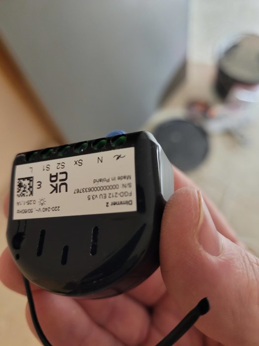 Hand håller en Schneider Exxact Primo dimmer med synliga anslutningsterminaler, suddig bakgrund med inredningsdetaljer.