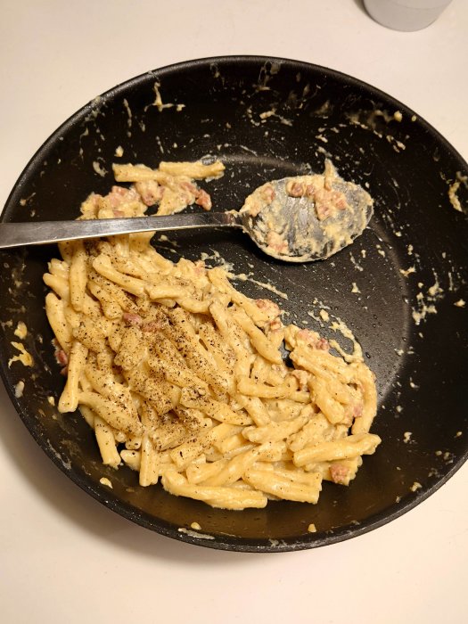 Hemlagad pasta carbonara i svart stekpanna med gaffel och tillbringare i bakgrunden