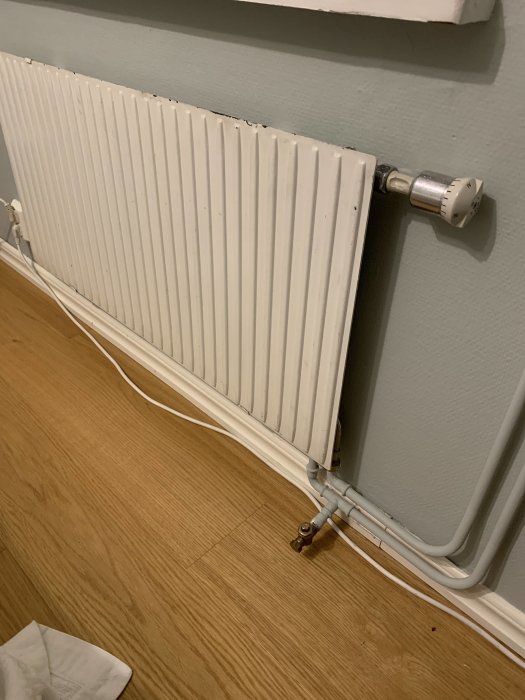 Vit radiator med termostat i öppet läge och en ovanlig ventil på returröret mot trägolv.