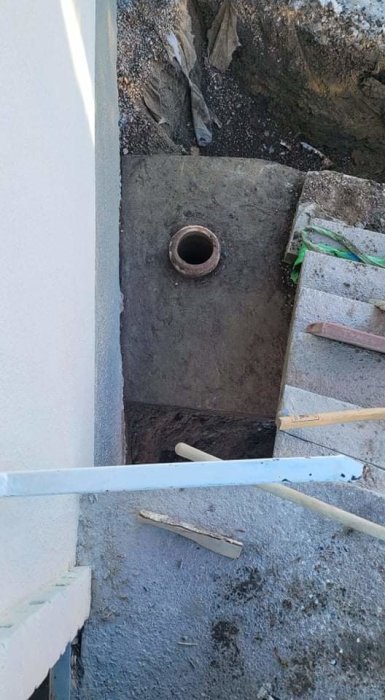 Stor betongpelare med hål grävd vid husdränering, nära källarvägg under trapp.