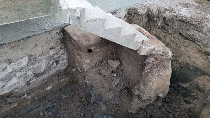 En utgrävd betongkonstruktion med ett stort hål vid en husgrund, synlig vid dräneringsarbete.