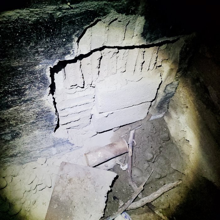 En kladdig källarvägg med sprickor och en igenmurad öppning, dämpad belysning, rubble och avfall på golvet.