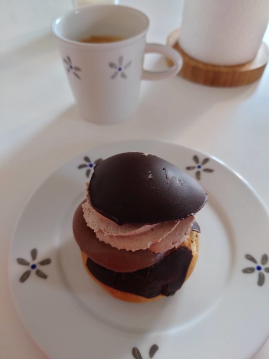 Svensk semla med mörk choklad på ett vitt fat, kaffekopp och servetthållare i bakgrunden.